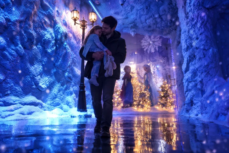 A dad carries his sleeping daughter back through the glowing ice corridor of the Ice Princess Suite at Electronic Ice Castle