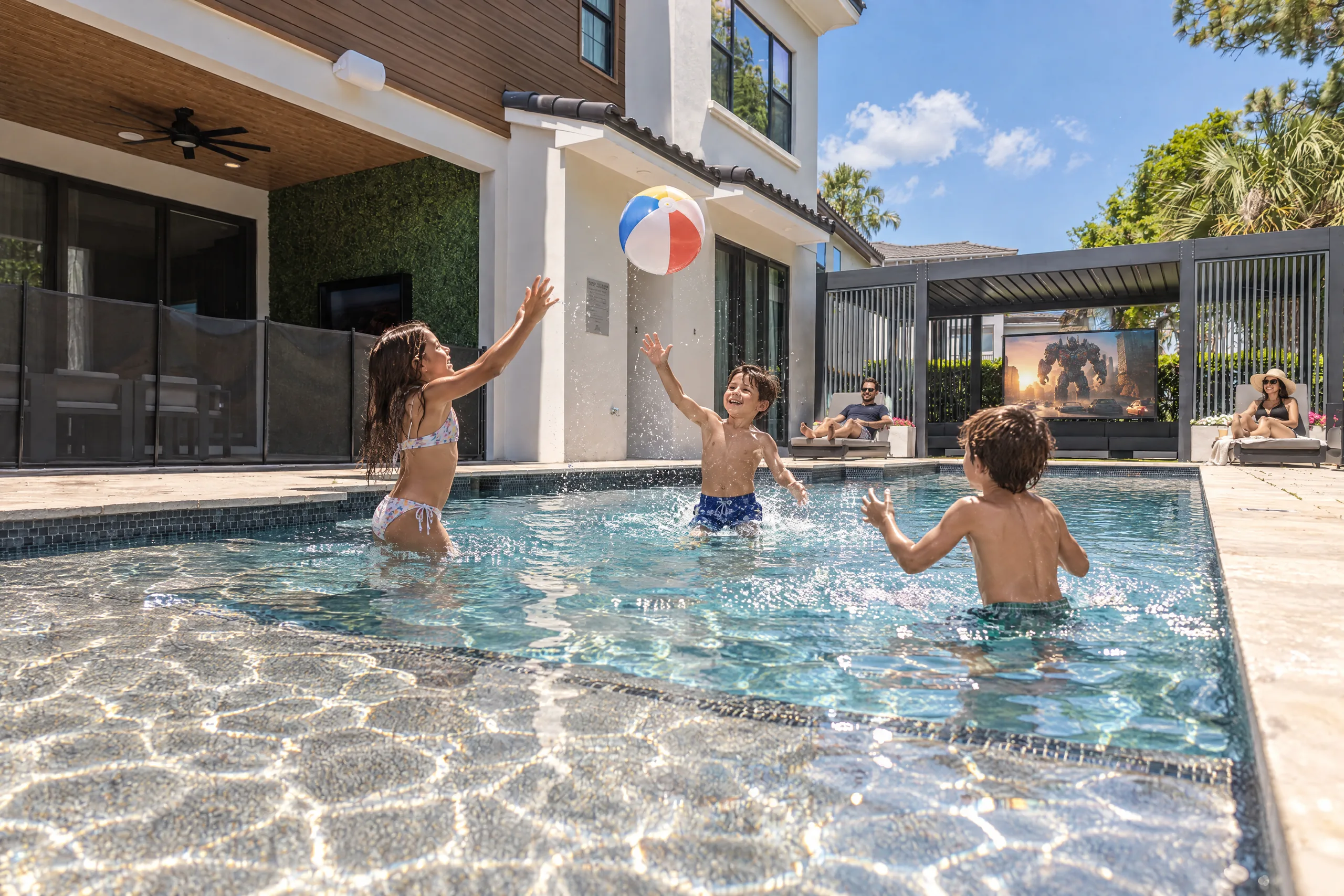 Kids toss a beach ball in the sunny Poolside Vibes pool while parents relax on loungers at The Iron Mansion